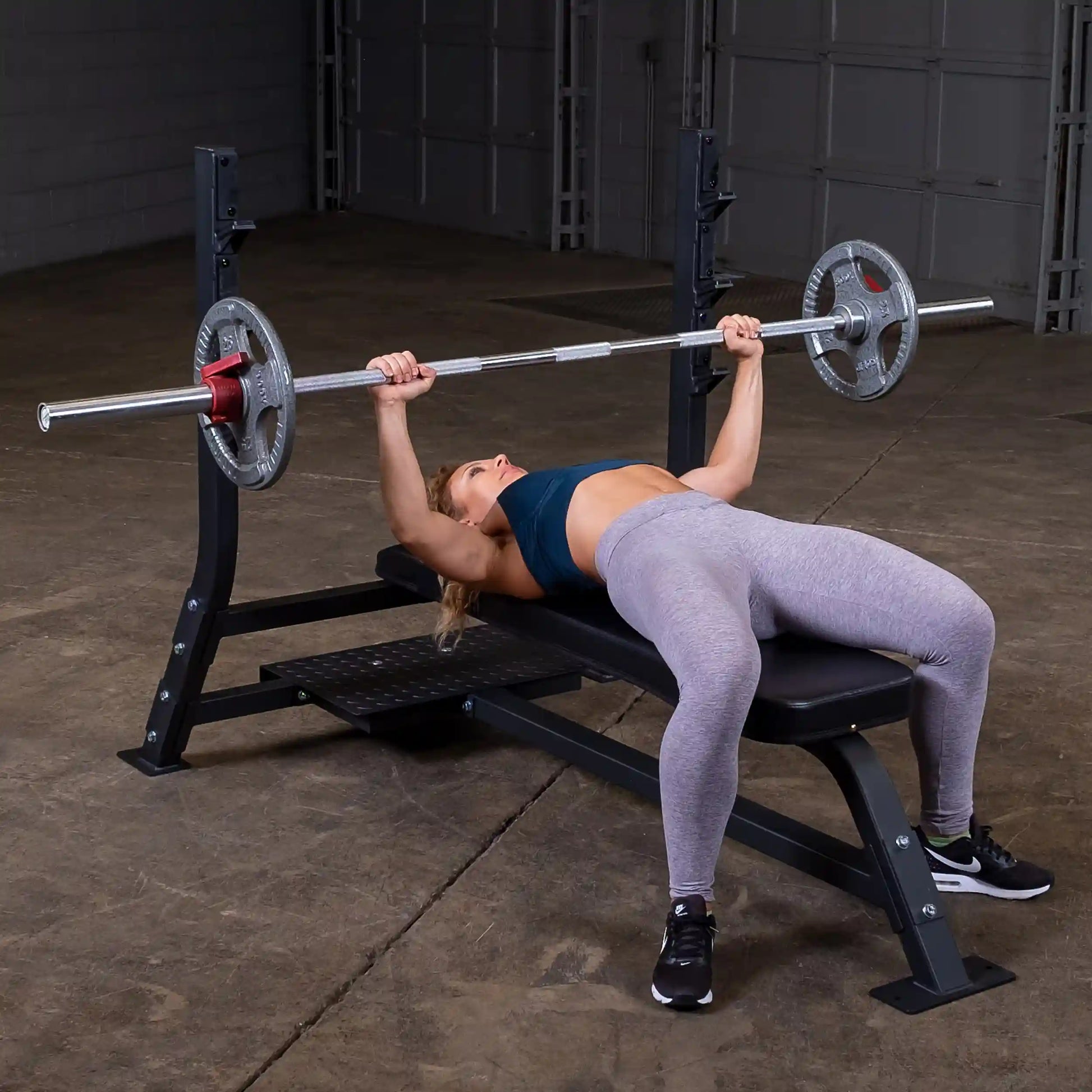 A woman performing a bench press on the Body-Solid Pro Clubline Flat Olympic Bench SOFB250, with a barbell and weights, in a gym setting.