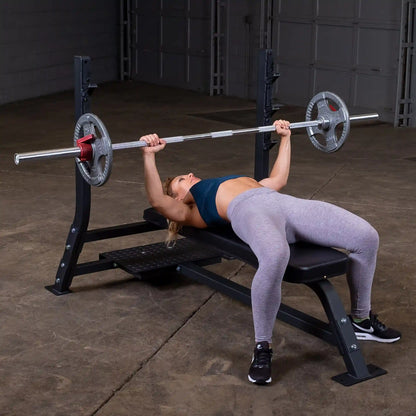 A woman performing a bench press on the Body-Solid Pro Clubline Flat Olympic Bench SOFB250, with a barbell and weights, in a gym setting.