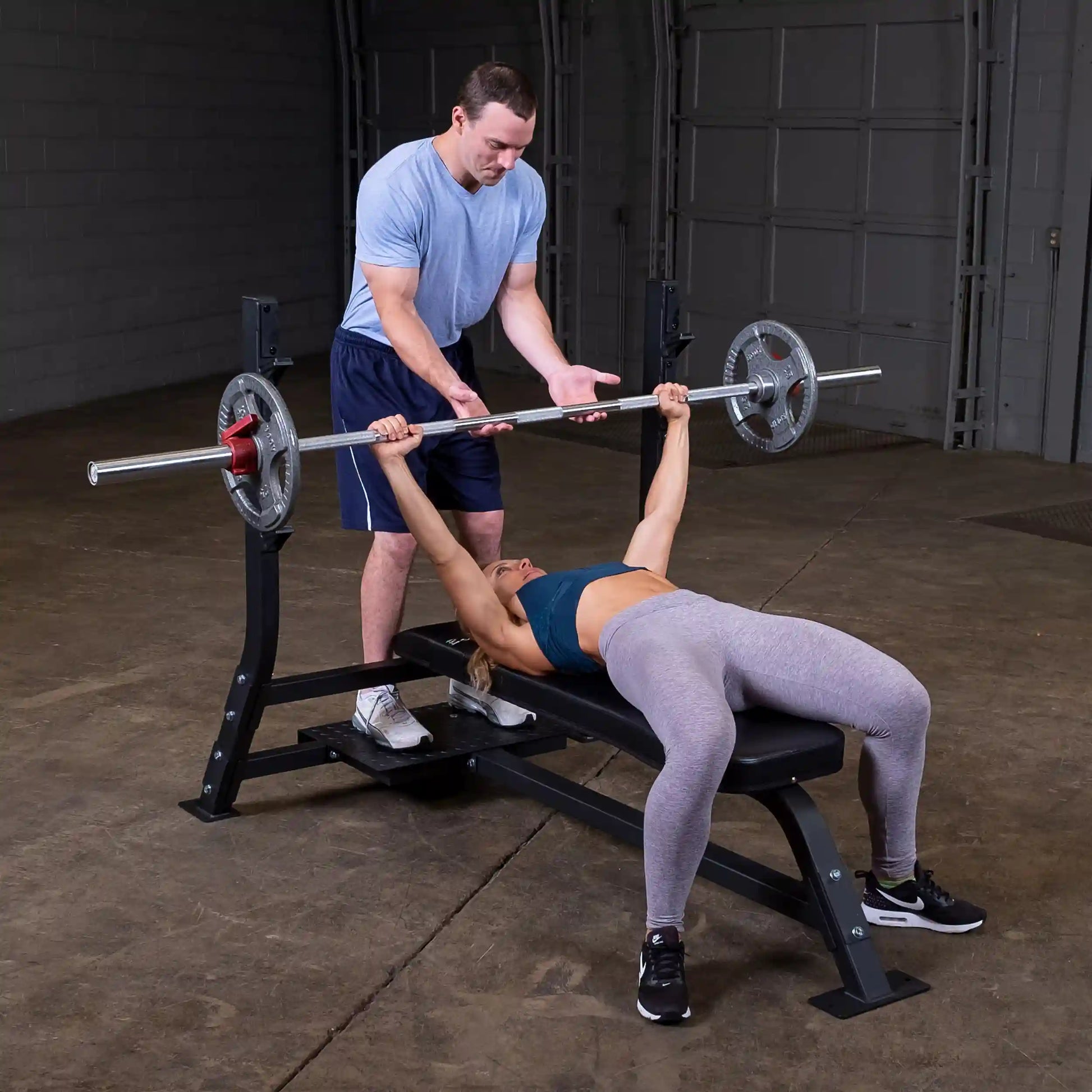 A man assists a woman as she performs a bench press on the Body-Solid Pro Clubline Flat Olympic Bench SOFB250 in a gym setting.