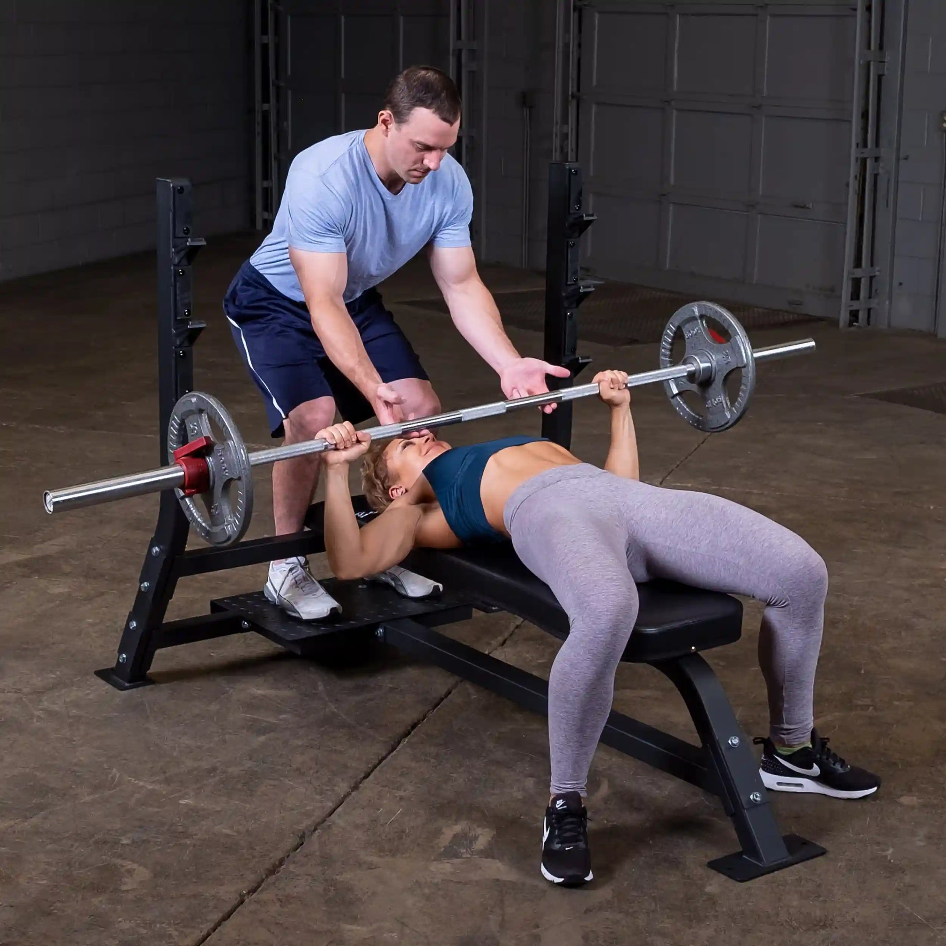 A man assisting a woman as she performs a bench press on the Body-Solid Pro Clubline Flat Olympic Bench SOFB250, with weights on the barbell in a gym setting.