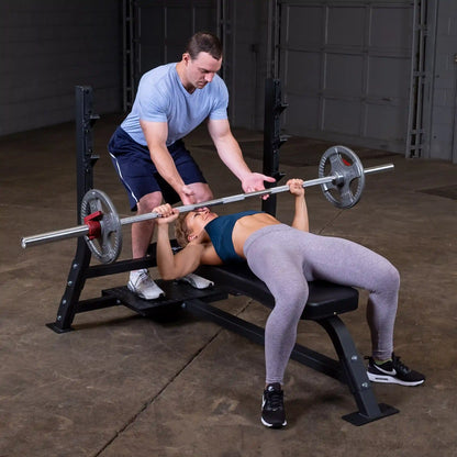 A man assisting a woman as she performs a bench press on the Body-Solid Pro Clubline Flat Olympic Bench SOFB250, with weights on the barbell in a gym setting.