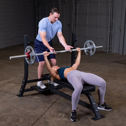 A man assists a woman lifting a barbell on the Body-Solid Pro Clubline Flat Olympic Bench SOFB250 in a gym setting.