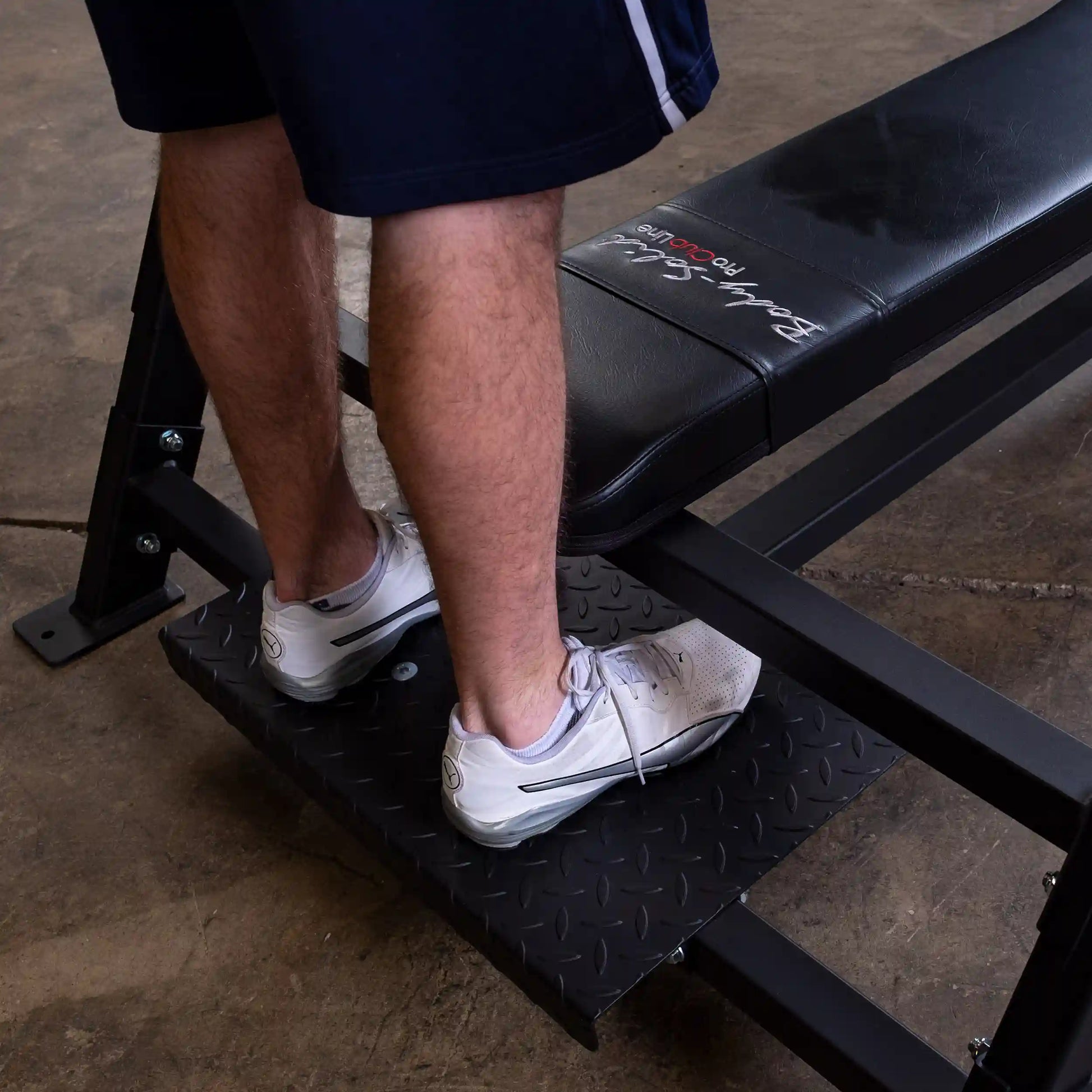 A person's feet on the footrest of the Body-Solid Pro Clubline Flat Olympic Bench SOFB250, showcasing the sturdy design and black finish of the equipment.