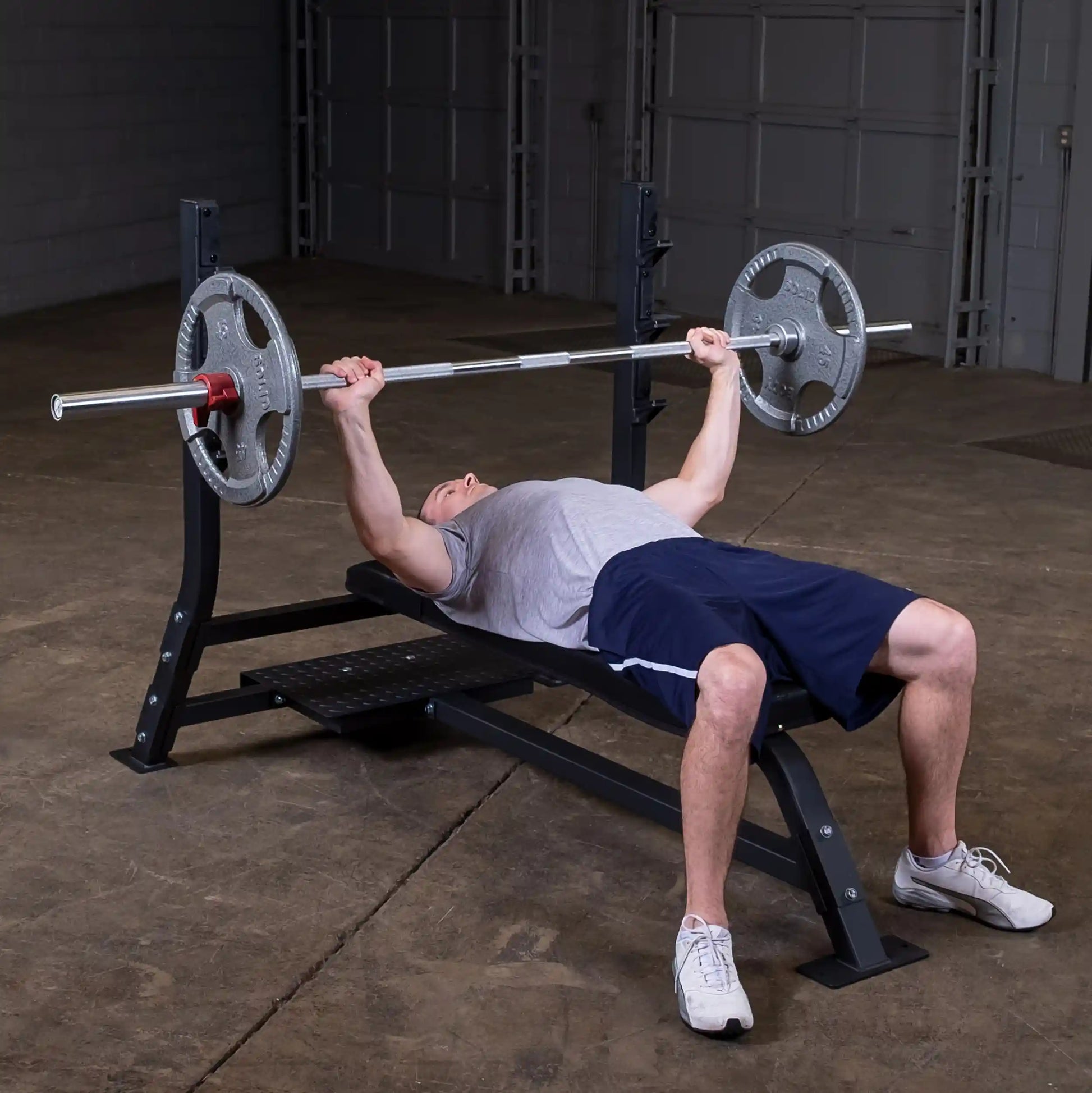 A man performing a bench press on the Body-Solid Pro Clubline Flat Olympic Bench SOFB250, with a barbell loaded with weights, in a gym setting.