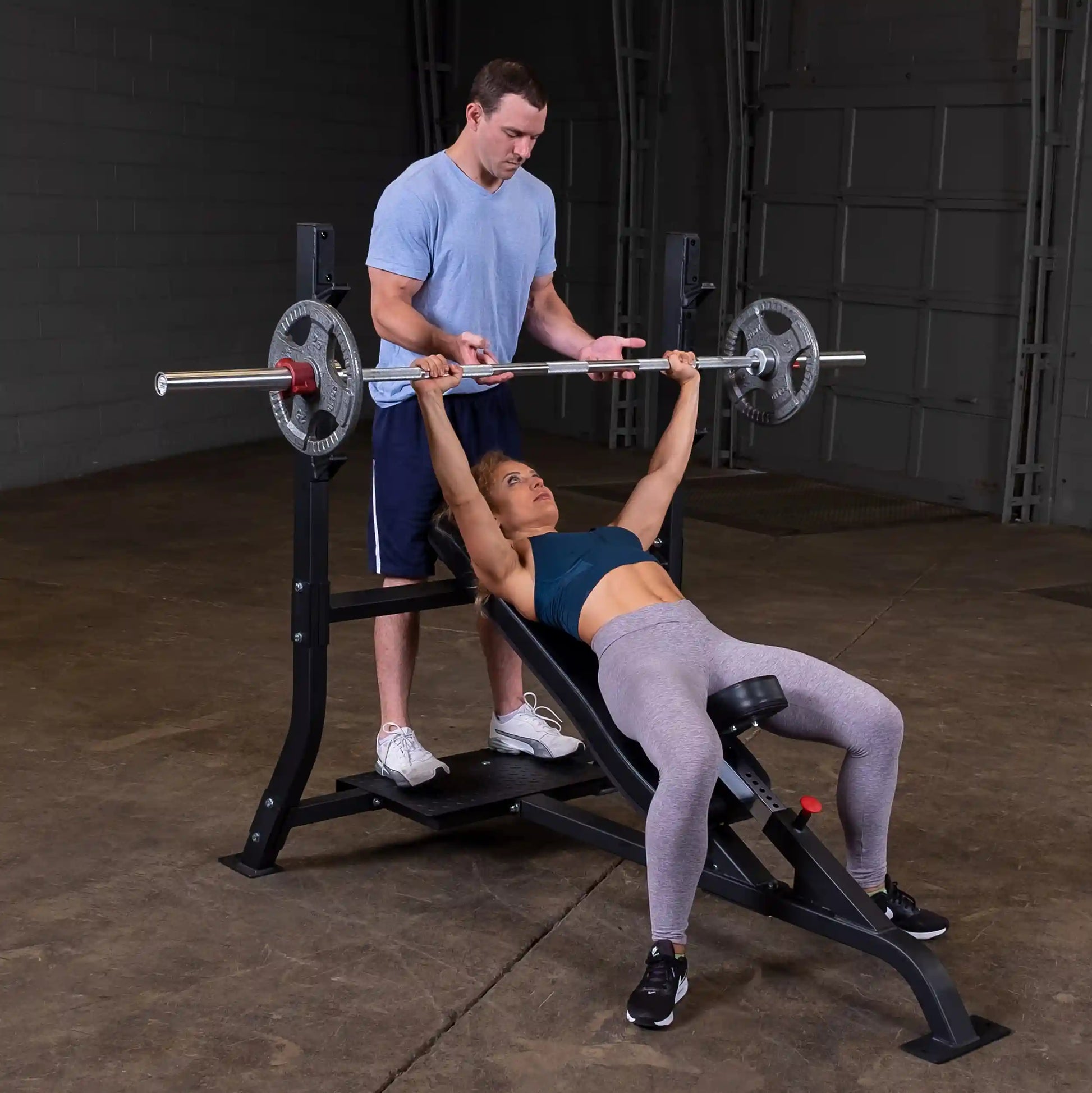 A woman performing an incline bench press with the assistance of a man, using the Body-Solid Pro Clubline Incline Olympic Bench SOIB250 in a gym setting.
