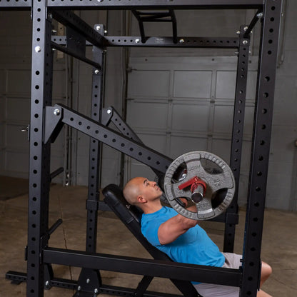 A person using the Body-Solid Pro Clubline Jammer Arms SPRJAM in a fitness setting, performing an exercise while lying on a bench beneath a weight rack.