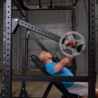 A man performing an overhead press on a bench using the Body-Solid Pro Clubline Jammer Arms SPRJAM, with a weight plate on the bar, in a gym setting with a rack in the background.