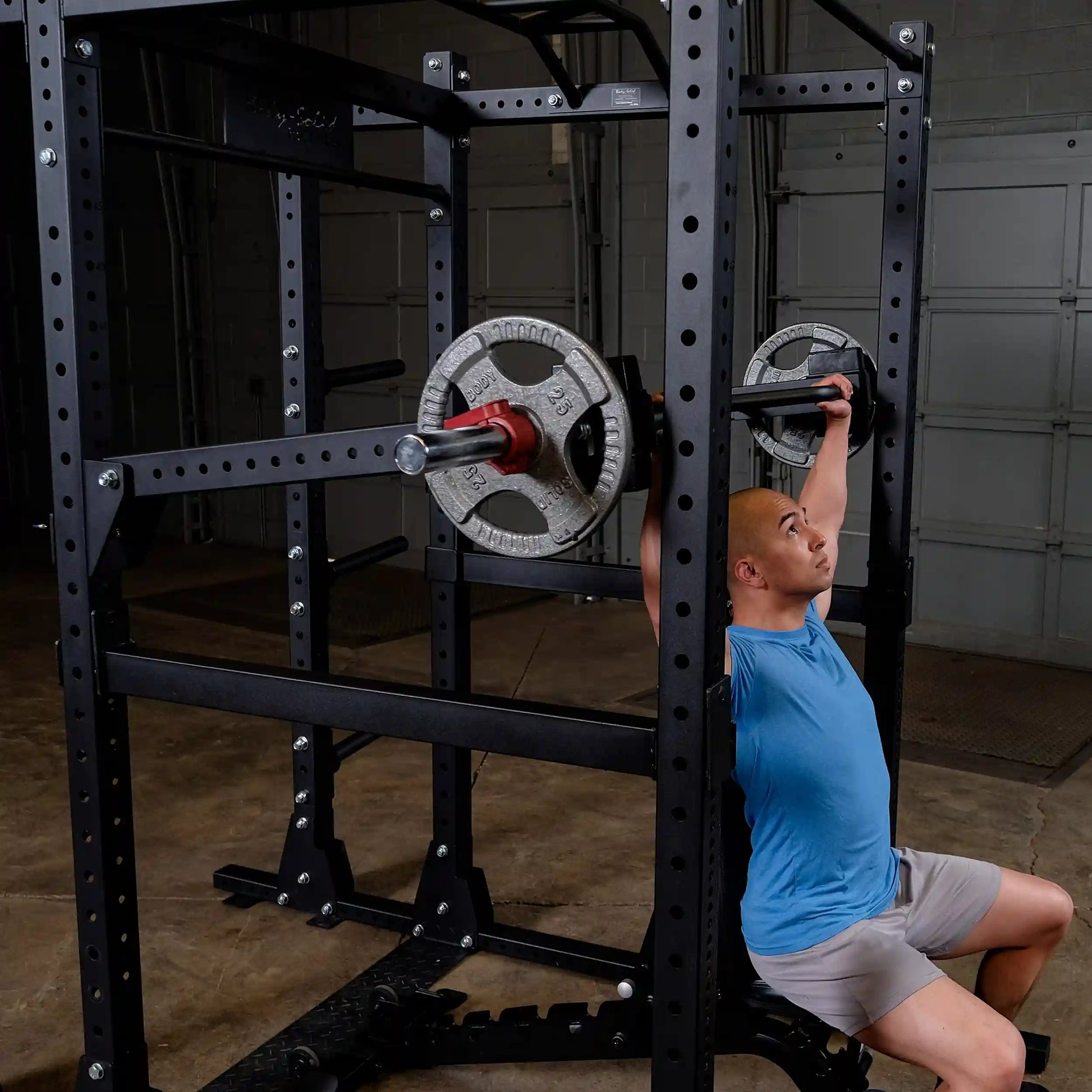A person using the Body-Solid Pro Clubline Jammer Arms SPRJAM for an overhead press workout in a gym setting, featuring a weightlifting rack with plates.