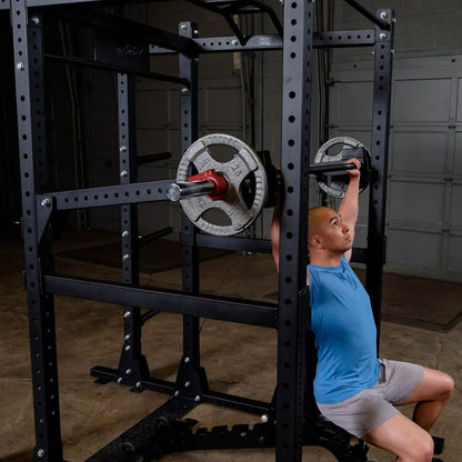 A person using the Body-Solid Pro Clubline Jammer Arms SPRJAM for an overhead press workout in a gym setting, featuring a weightlifting rack with plates.