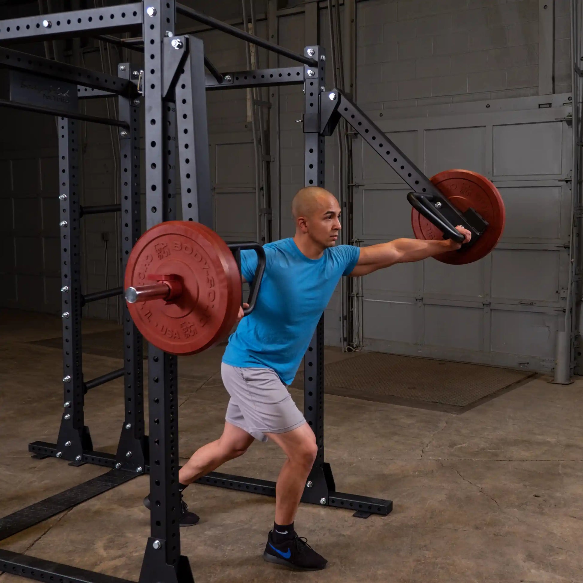 A man demonstrating an exercise using the Body-Solid Pro Clubline Jammer Arms SPRJAM, with a barbell and weights, in a fitness gym setting.