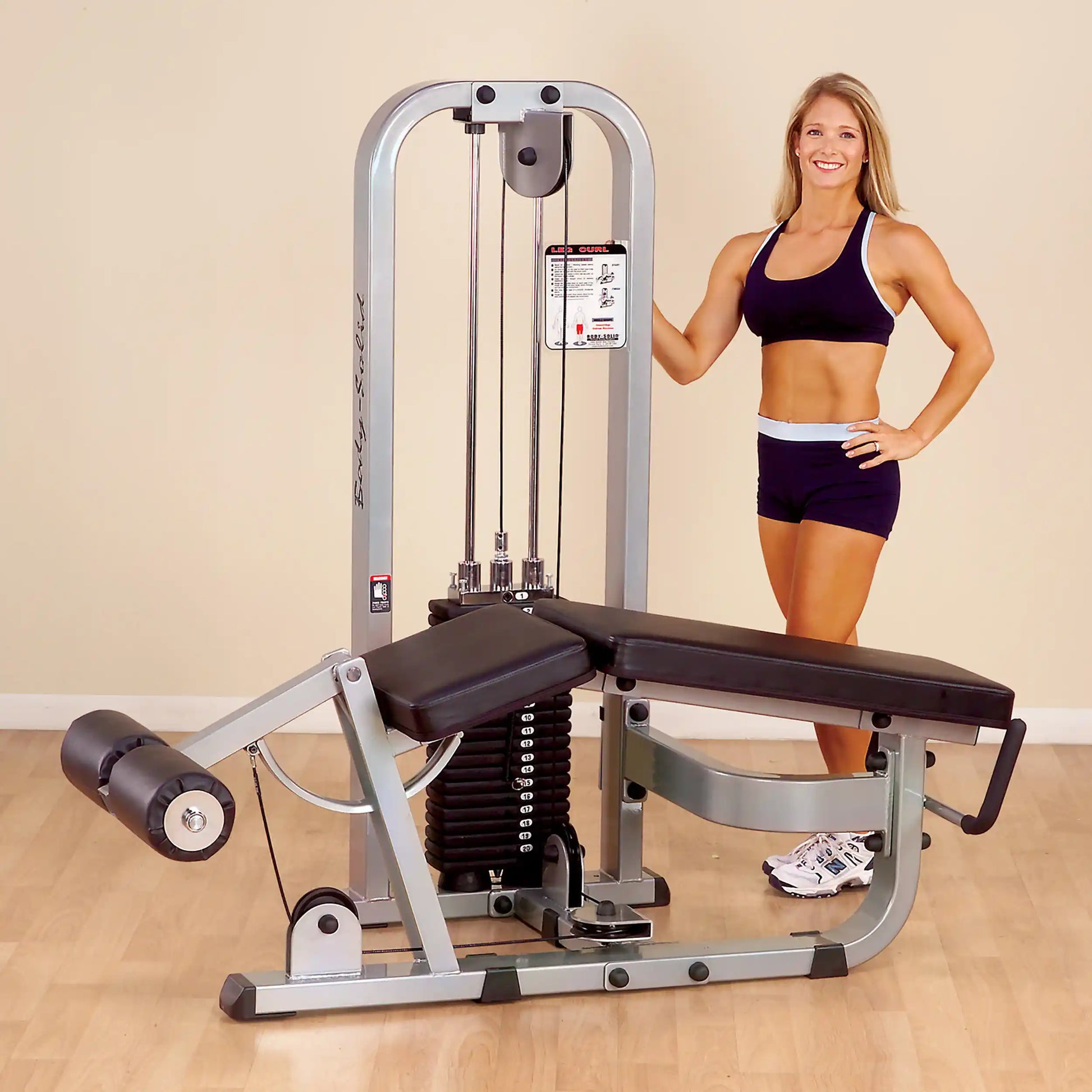 A fitness model stands next to the Body-Solid Pro Clubline Leg Curl Machine SLC400G, showcasing its design and features in a gym setting.