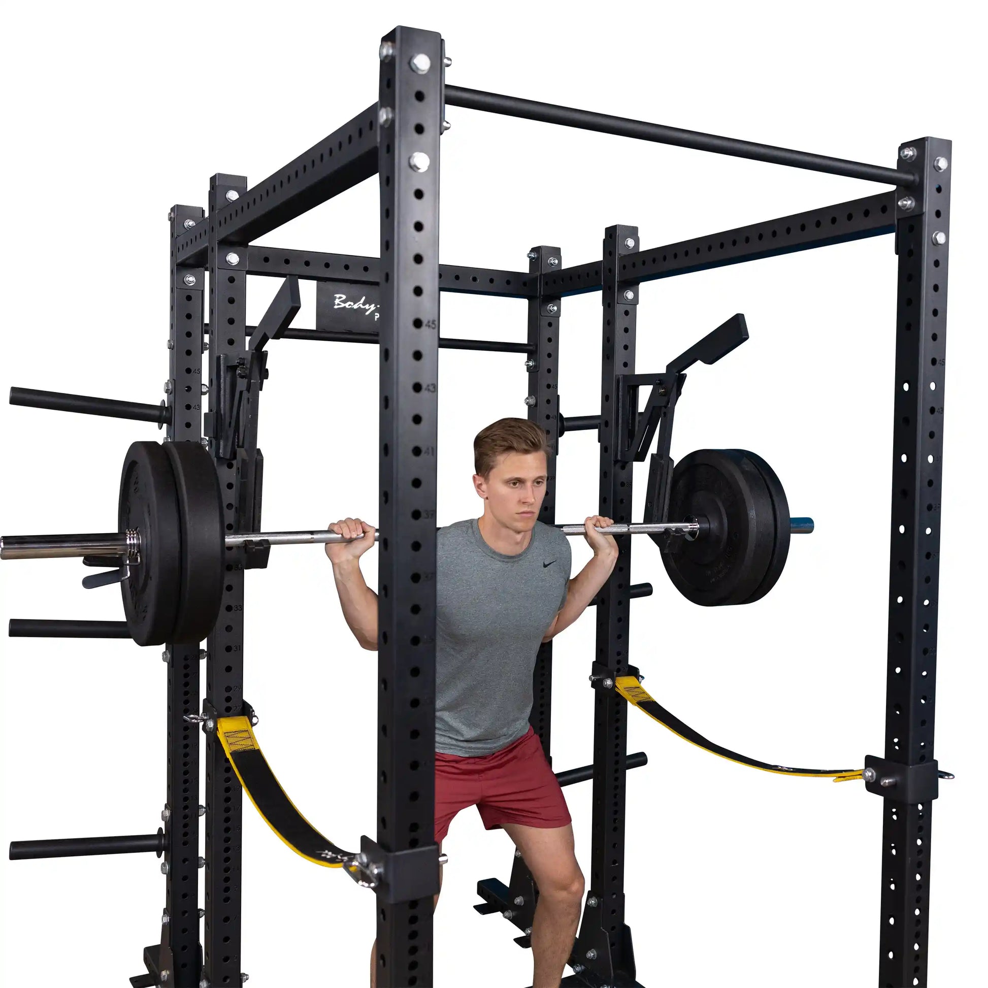 A person performing a squat with a loaded barbell in a Body-Solid Pro Clubline Monolift Attachment SPRLFT, featuring a sturdy black power rack with safety straps and weight plates.