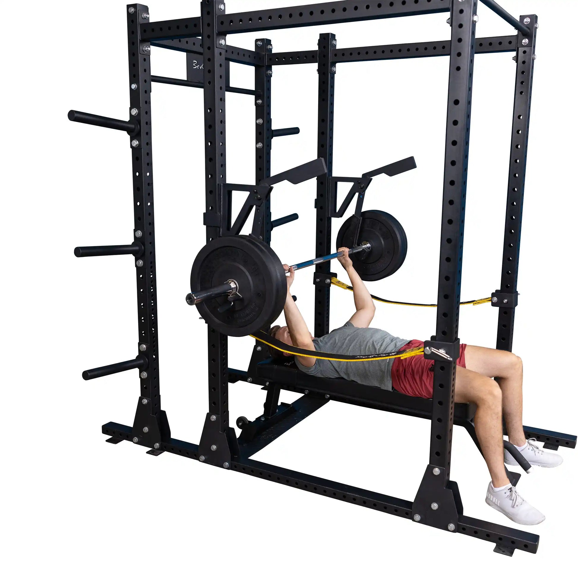 A person performing a bench press using the Body-Solid Pro Clubline Monolift Attachment SPRLFT, featuring a black power rack, weight plates, and safety bands.