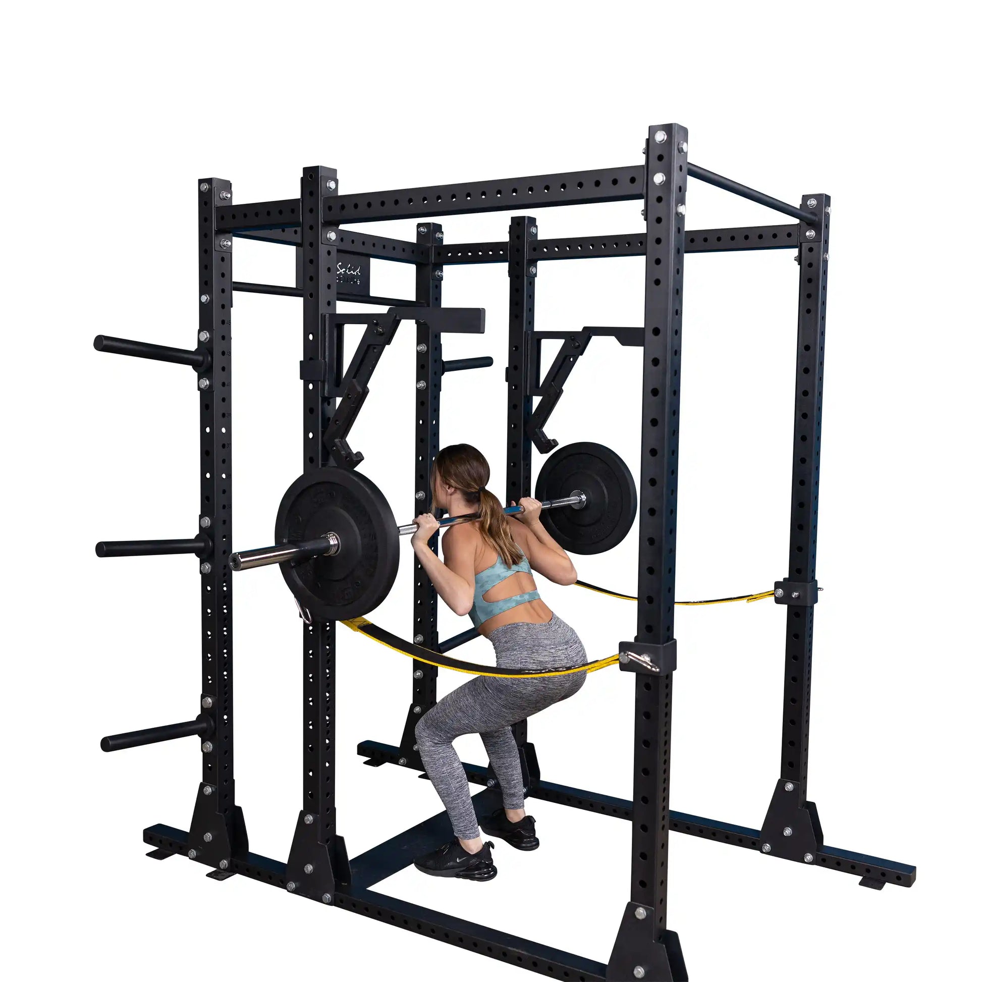 A person using the Body-Solid Pro Clubline Monolift Attachment SPRLFT to perform squats with a barbell in a gym setting. The equipment features a sturdy black frame with weight plates and safety features for secure lifting.