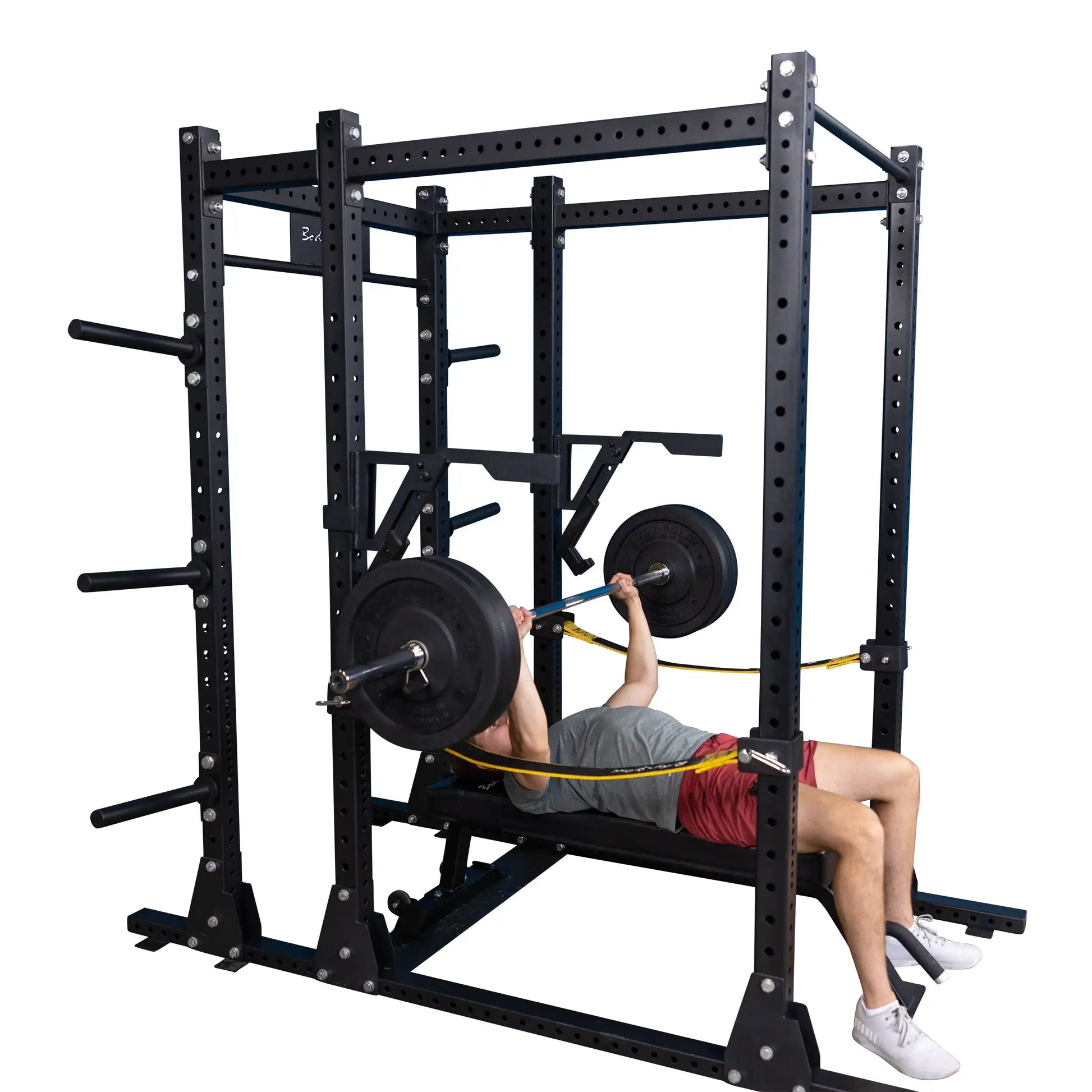 A person using the Body-Solid Pro Clubline Monolift Attachment SPRLFT to perform a bench press in a squat rack, featuring black metal framing and weight plates.