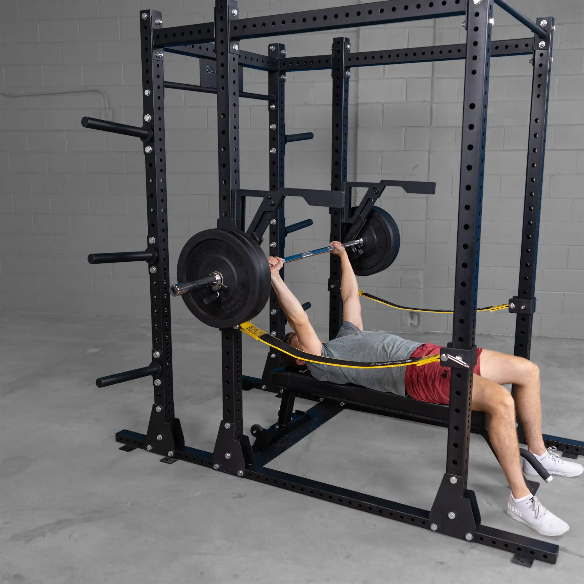 A person performing a bench press using the Body-Solid Pro Clubline Monolift Attachment SPRLFT, surrounded by a squat rack setup on a gym floor.