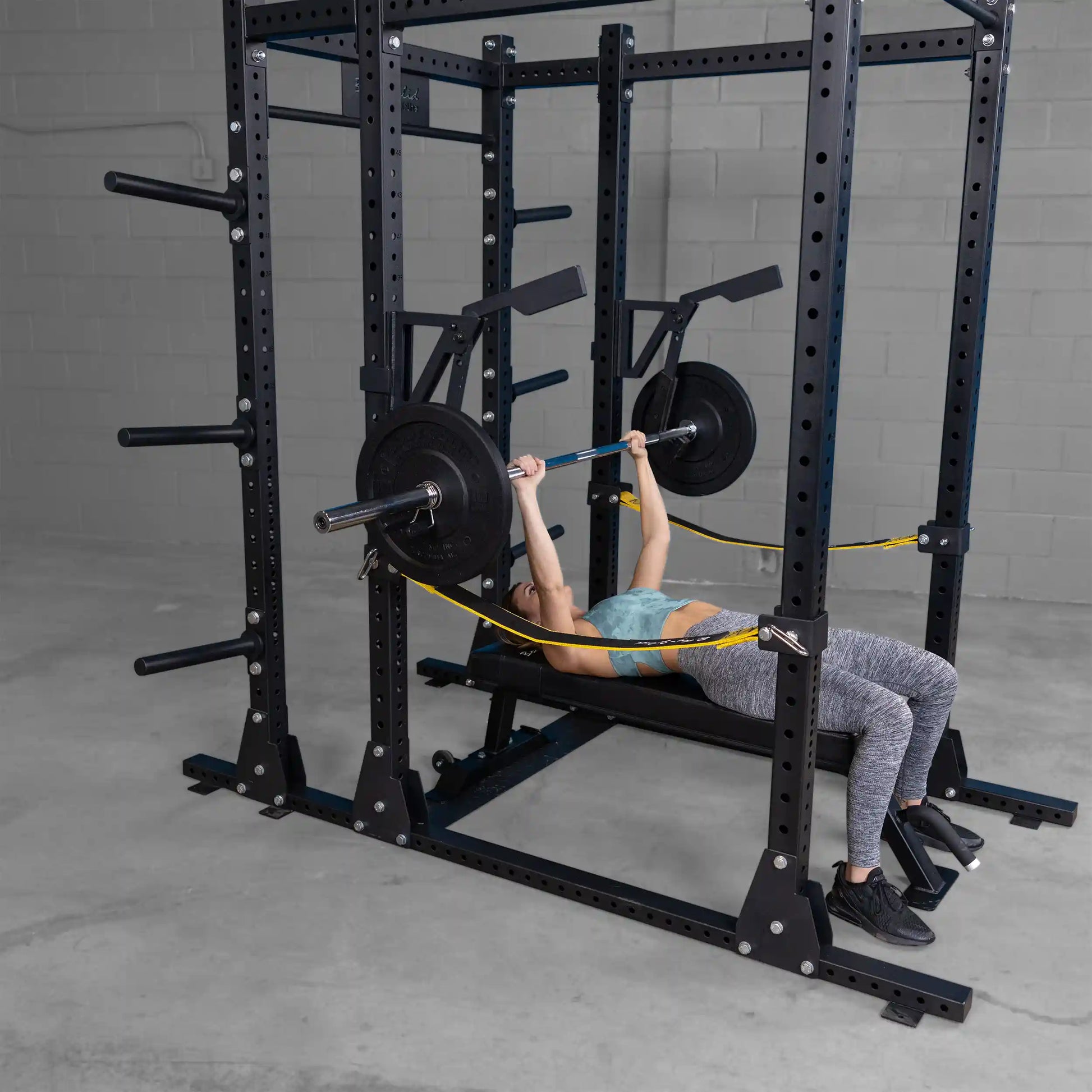A person performing a bench press with the Body-Solid Pro Clubline Monolift Attachment SPRLFT in a gym setting, featuring a squat rack with weight plates and resistance bands.