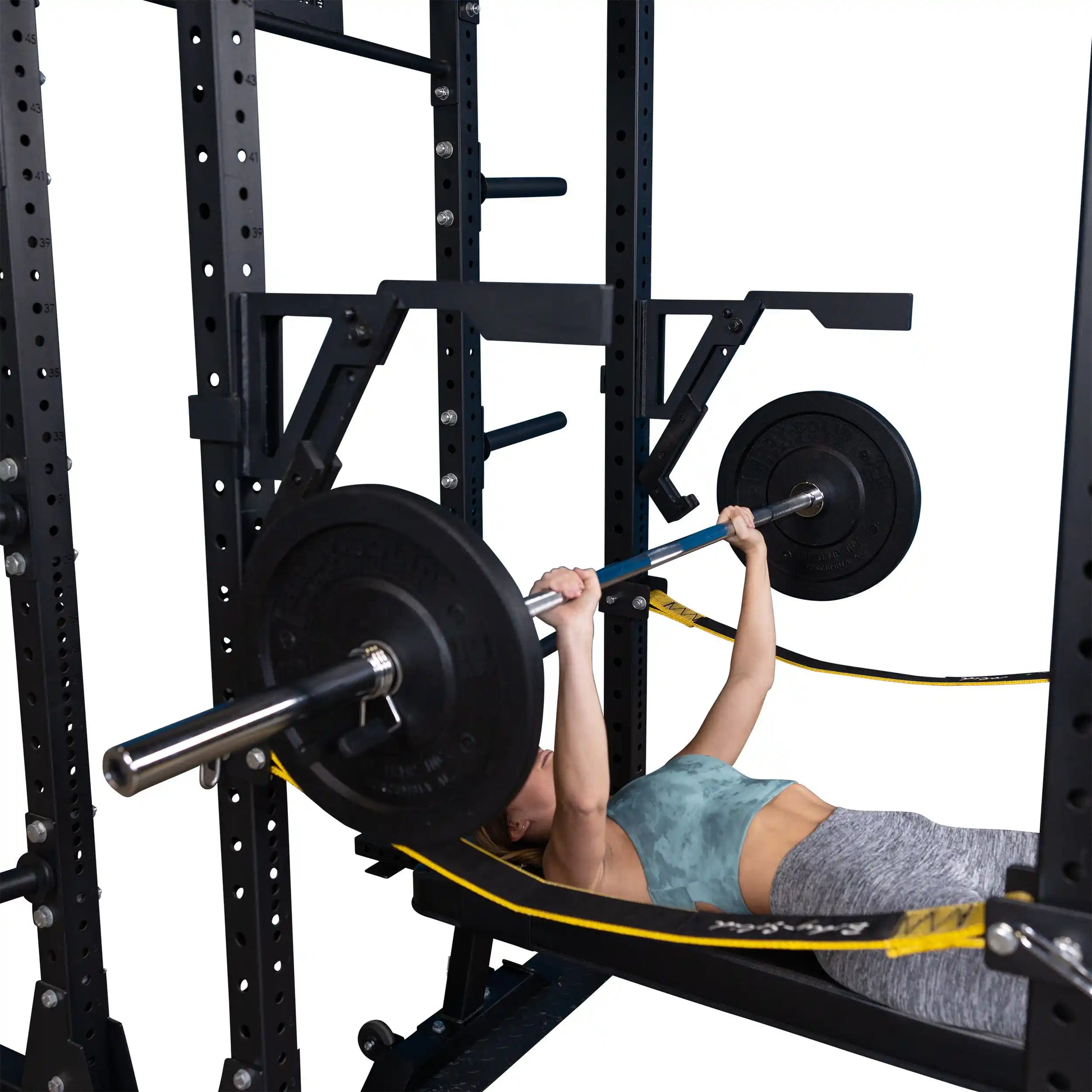 A person performing a bench press using the Body-Solid Pro Clubline Monolift Attachment SPRLFT, with a barbell and weights. The setup includes a squat rack in the background and resistance bands attached.