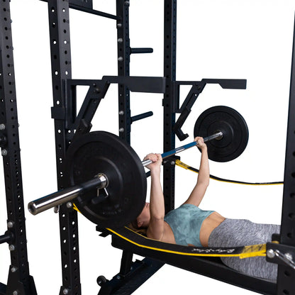 A person performing a bench press using the Body-Solid Pro Clubline Monolift Attachment SPRLFT, with a barbell and weights. The setup includes a squat rack in the background and resistance bands attached.