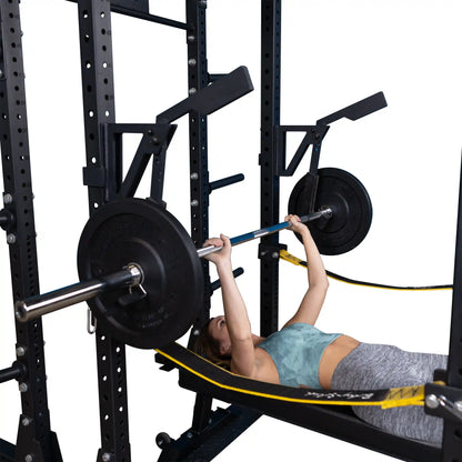 A person performing a bench press using the Body-Solid Pro Clubline Monolift Attachment SPRLFT, with a barbell loaded with weight plates and a sturdy power rack in the background.