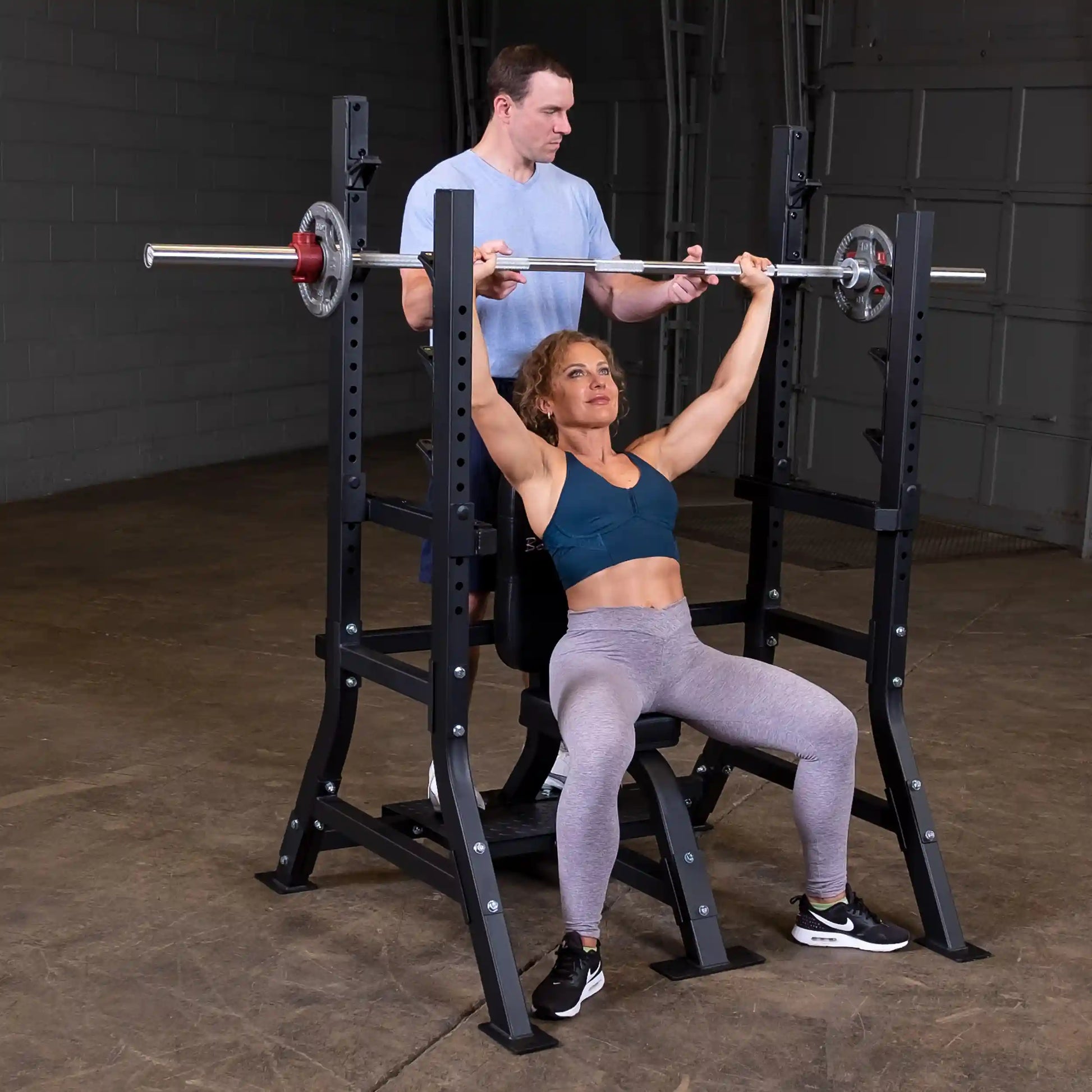 A woman performs an overhead press on the Body-Solid Pro Clubline Shoulder Olympic Bench SOSB250 while a man assists her.