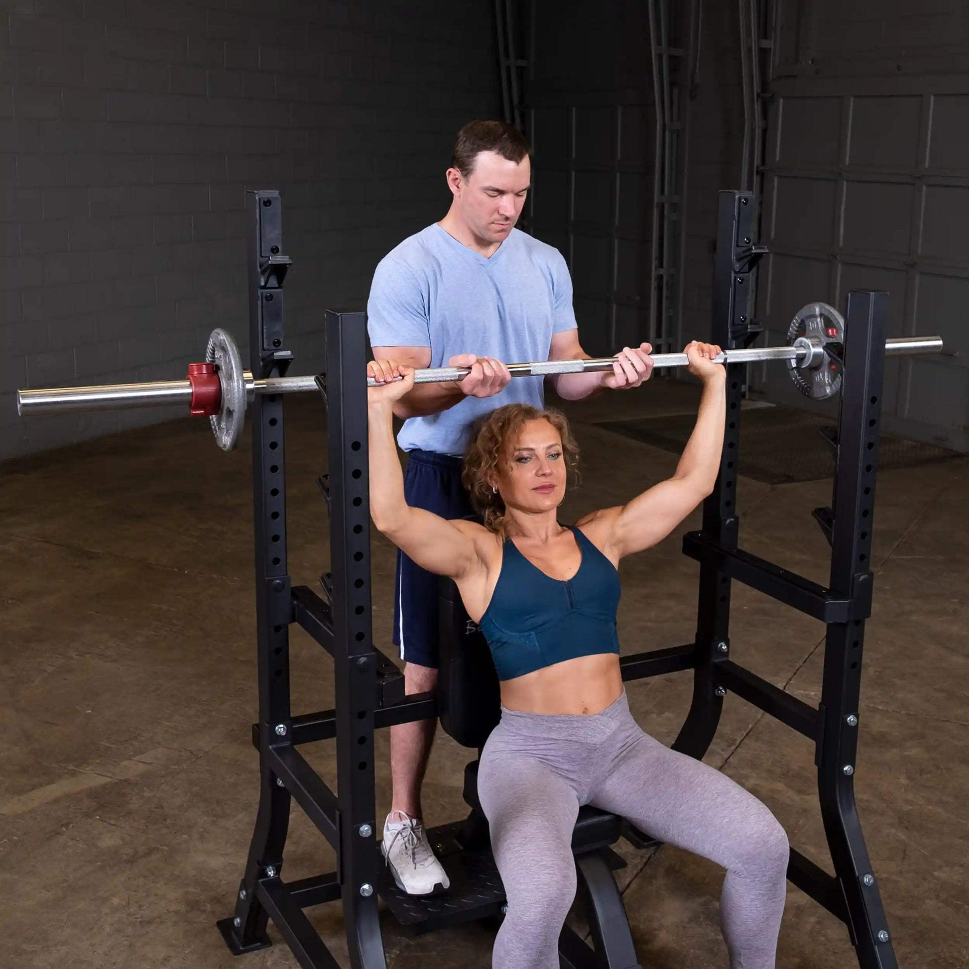 A woman performing an overhead press with a barbell while using the Body-Solid Pro Clubline Shoulder Olympic Bench SOSB250, assisted by a man standing behind her.