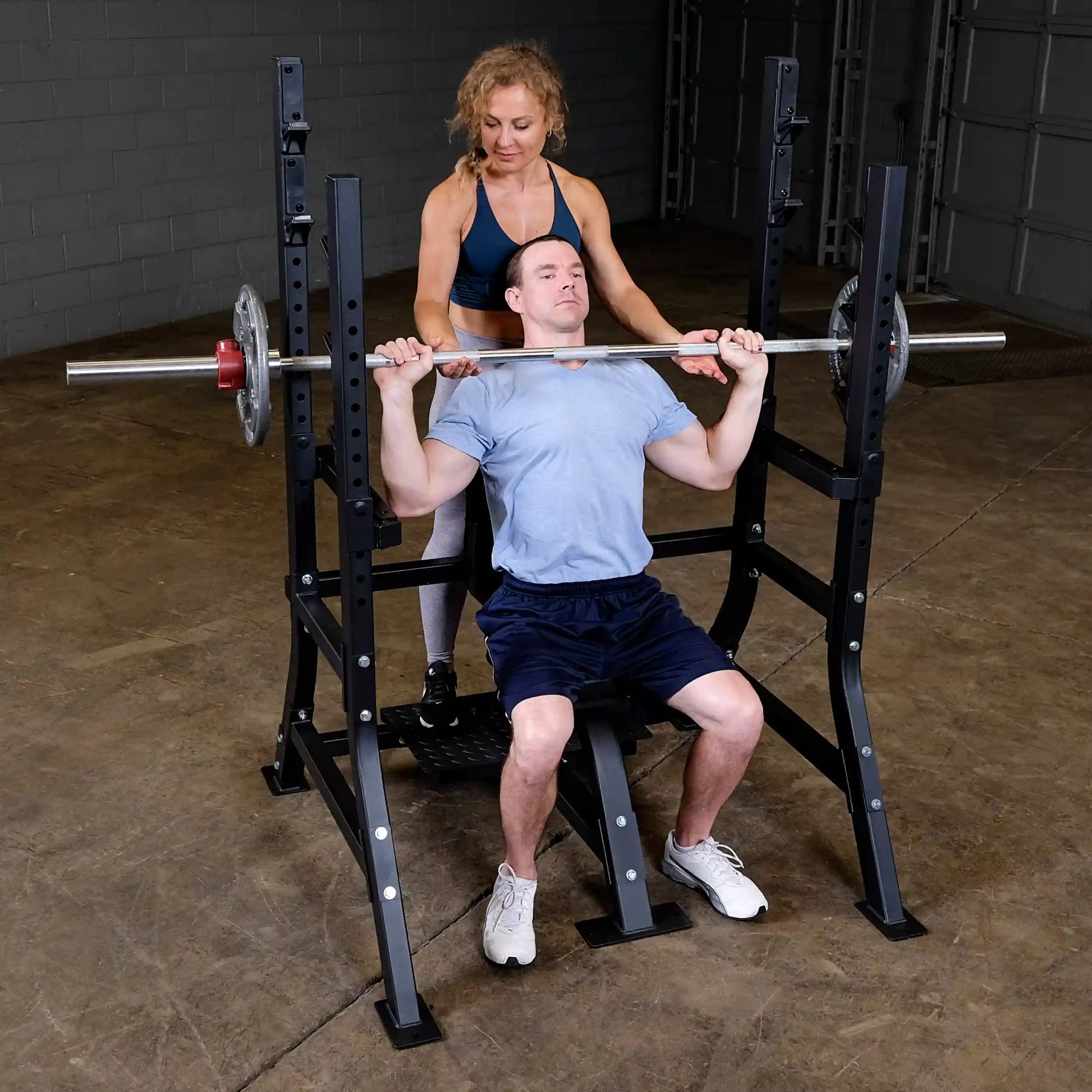 A man seated on the Body-Solid Pro Clubline Shoulder Olympic Bench SOSB250, performing a shoulder press with a barbell, while a woman assists him from behind.