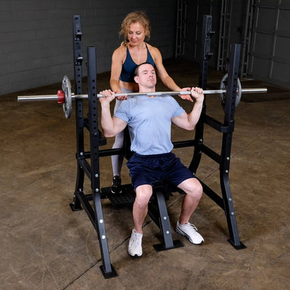 A man seated on the Body-Solid Pro Clubline Shoulder Olympic Bench SOSB250, performing a shoulder press with a barbell, while a woman assists him from behind.