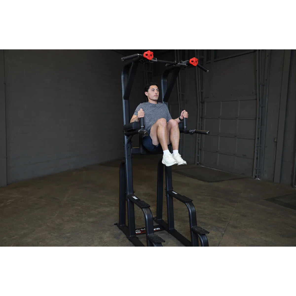 A person demonstrating the Body-Solid Pro Clubline Vertical Knee Raise SVKR1000 exercise equipment, seated with legs raised, in a gym setting.