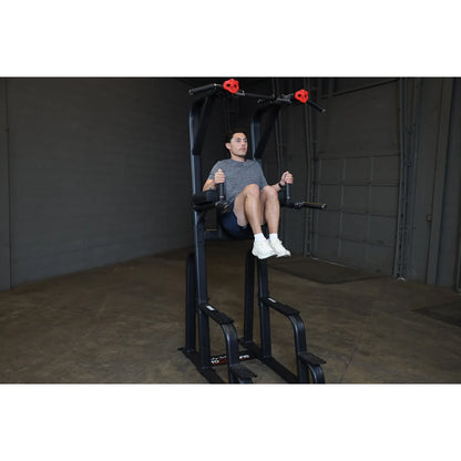 A person demonstrating the Body-Solid Pro Clubline Vertical Knee Raise SVKR1000 exercise equipment, seated with legs raised, in a gym setting.
