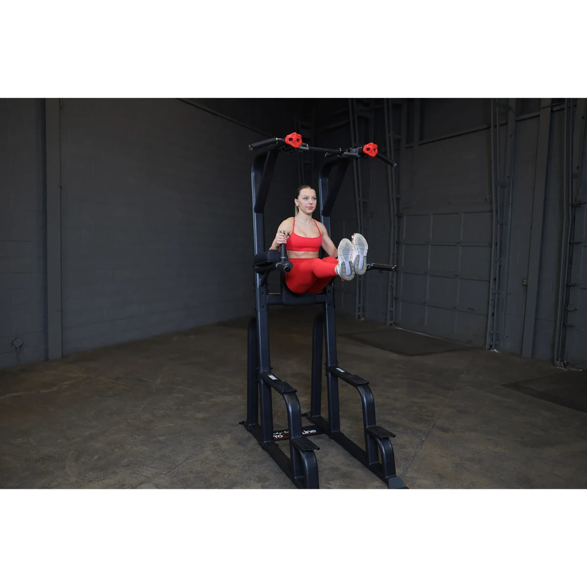 A woman in a red workout outfit demonstrates an exercise on the Body-Solid Pro Clubline Vertical Knee Raise SVKR1000, a fitness machine designed for core workouts, set in a gym environment.