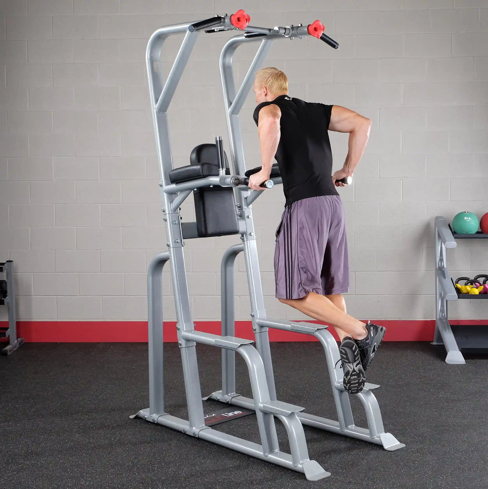 A man performing dips on the Body-Solid Pro Clubline Vertical Knee Raise SVKR1000 in a gym setting, showcasing strength training equipment with a gray and red color scheme.