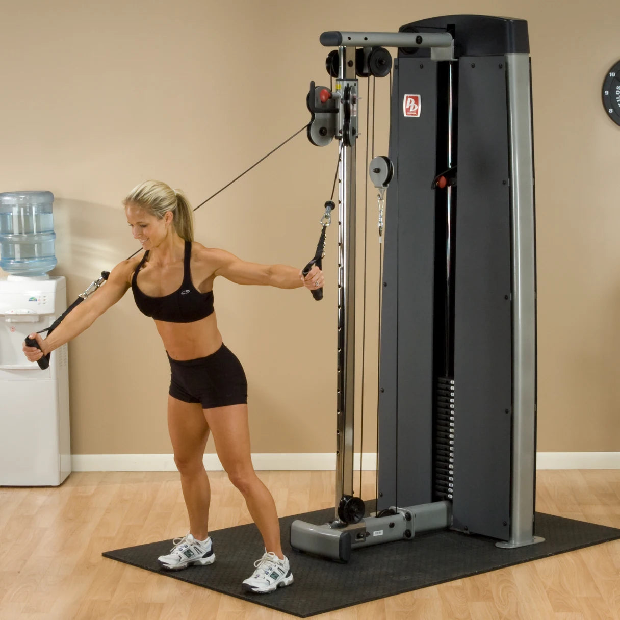 A woman performing an exercise using the Body-Solid Pro Dual Cable Column Machine DPCCSF in a gym setting, with a water cooler in the background.