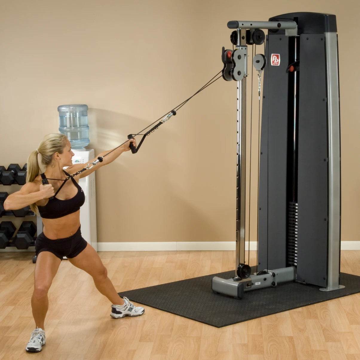 A woman performing an exercise using the Body-Solid Pro Dual Cable Column Machine DPCCSF in a gym setting, with dumbbells and a water cooler in the background.