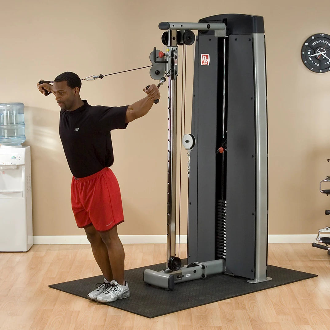 A man performing an exercise using the Body-Solid Pro Dual Cable Column Machine DPCCSF in a gym setting, with a water cooler and clock in the background.