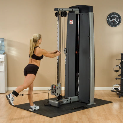 A woman exercises using the Body-Solid Pro Dual Cable Column Machine DPCCSF in a gym setting, demonstrating a cable workout with one leg extended behind her.