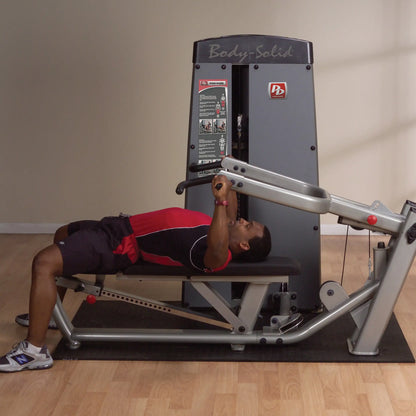 A person performing an exercise on the Body-Solid Pro Dual Multi-Press Machine DPRSSF, lying on a bench and gripping the machine's handles, in a fitness studio with wooden flooring.