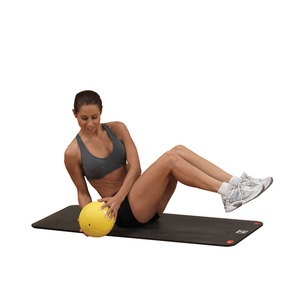 Woman performing an abdominal exercise on a black foam mat while holding a yellow medicine ball.