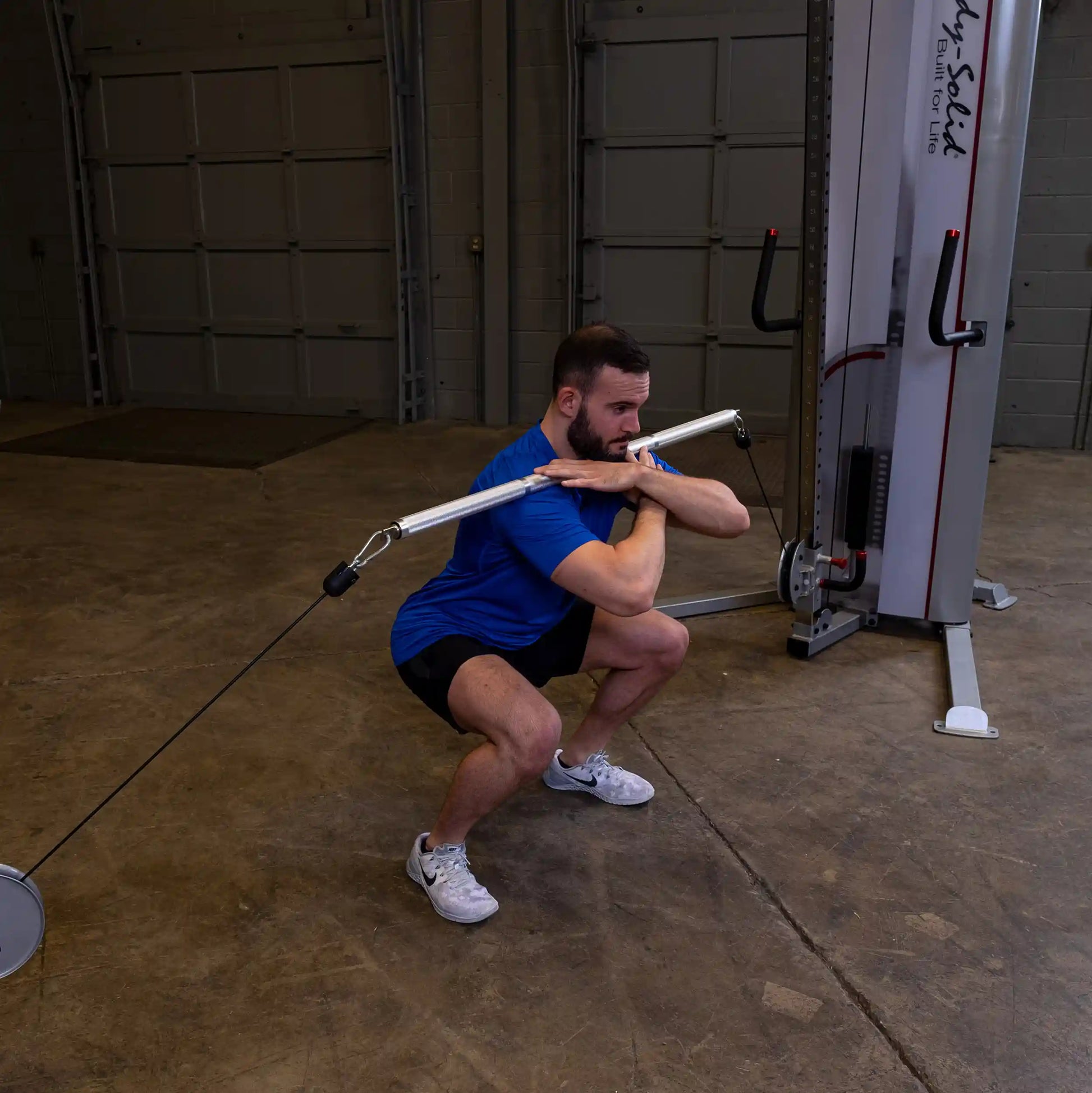 Man performing squats with Body-Solid functional trainer, wearing blue shirt and black shorts.