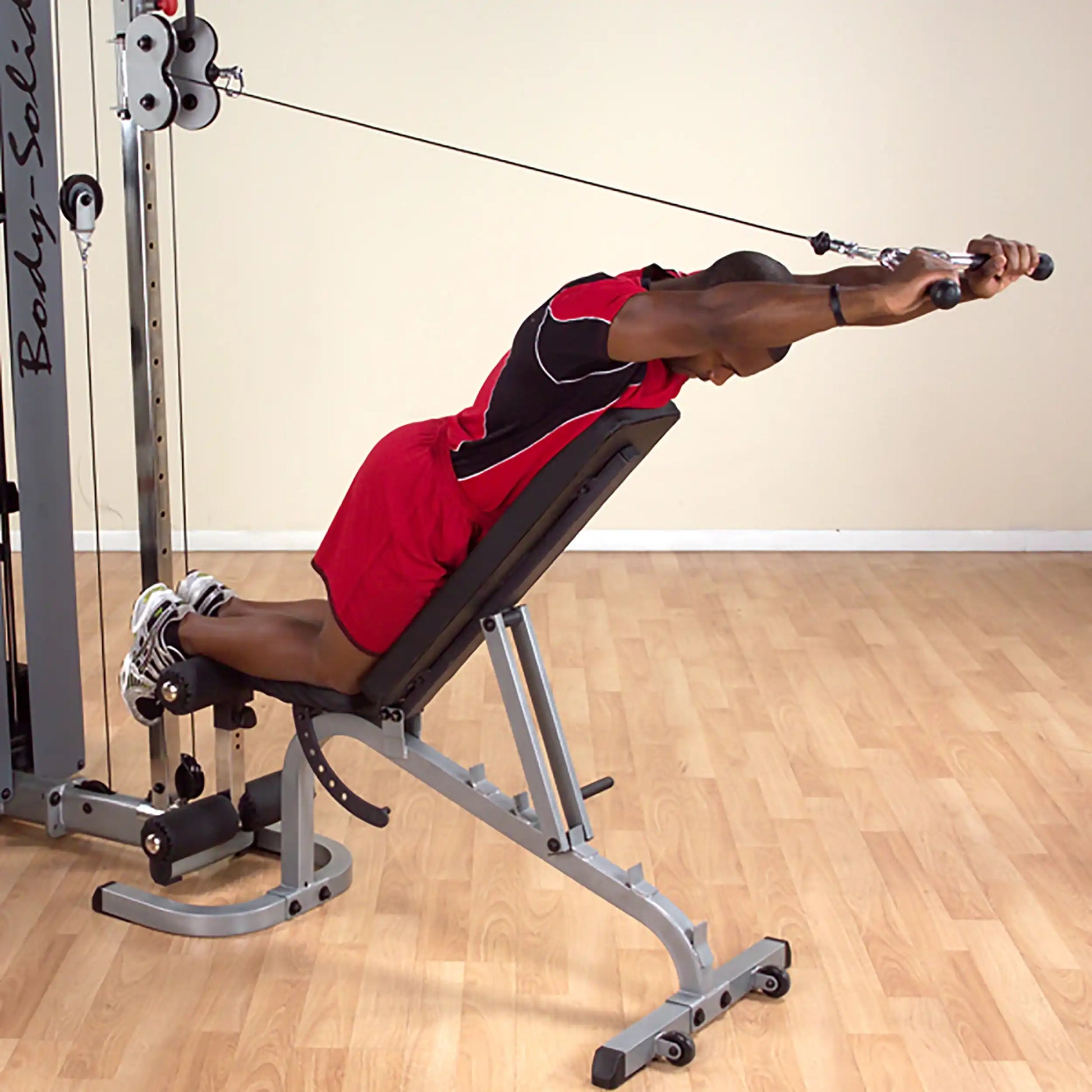 Man using Body-Solid cable machine for upper body workout on incline bench, wearing red athletic outfit.