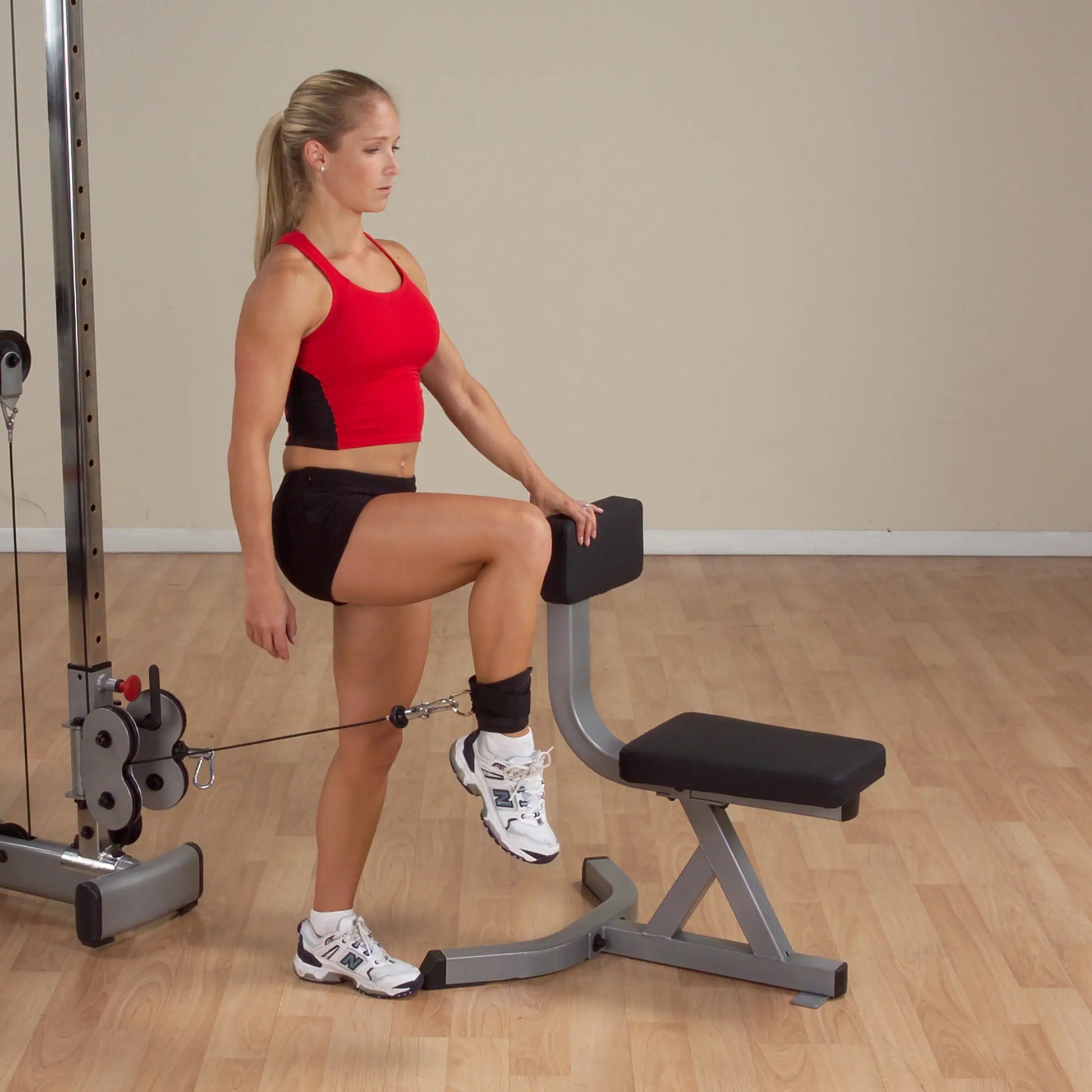 Woman using cable machine for leg exercise in a red top and black shorts.