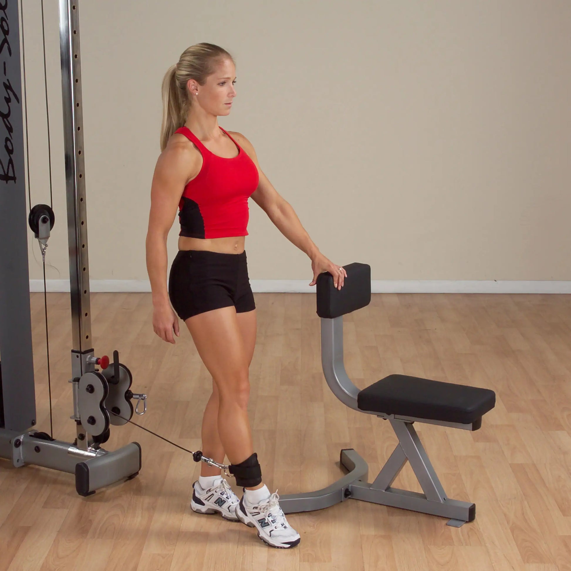 Woman in red tank top using Body-Solid cable machine for leg exercise.