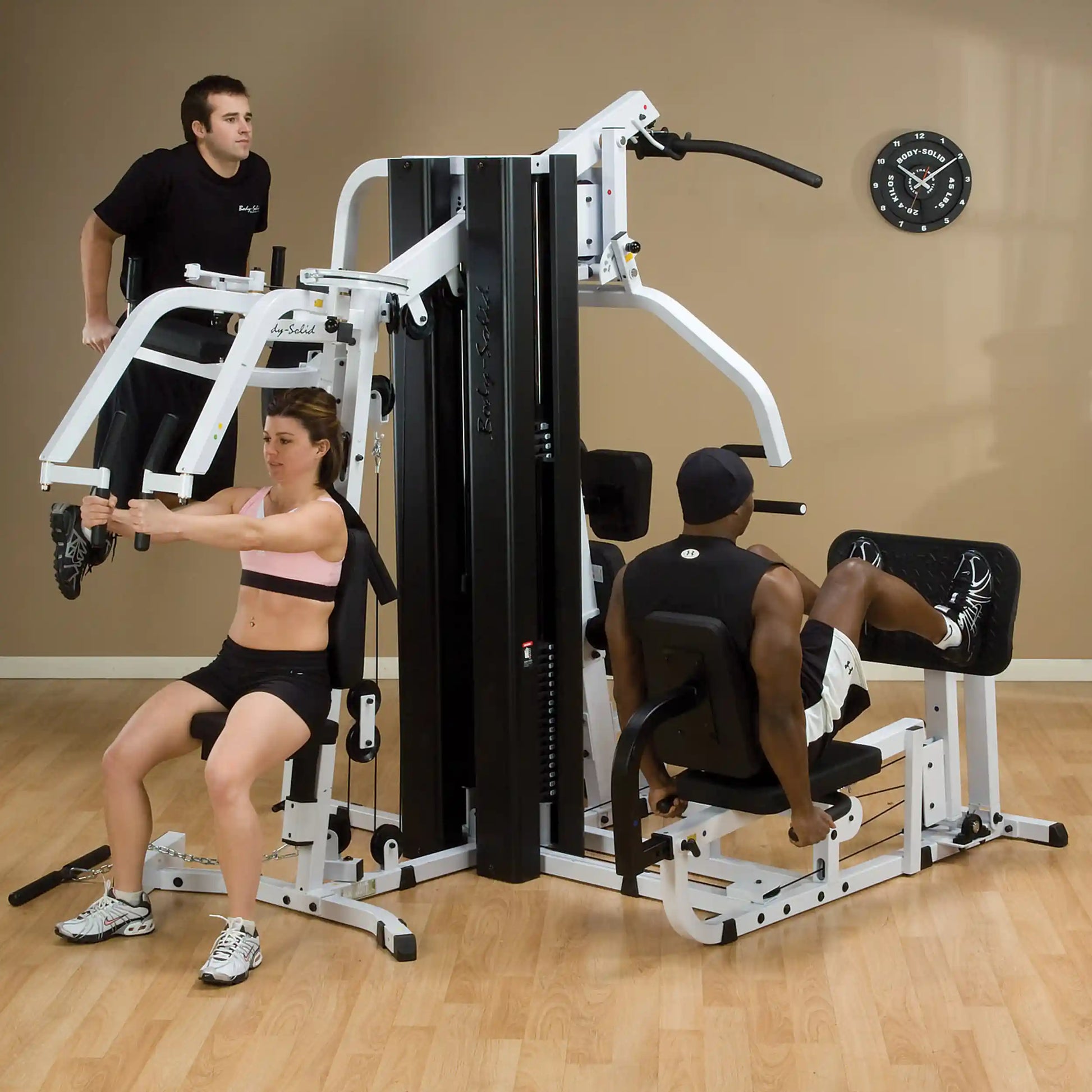 Body-Solid home gym with three people exercising on a multi-station machine in a fitness room.