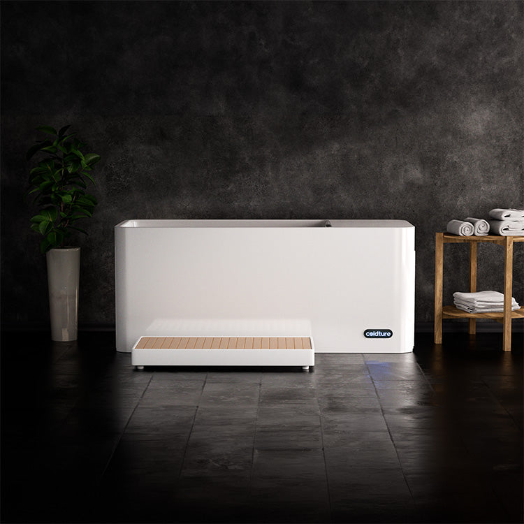 Coldture Pro Plunge ice bath setup featuring a sleek white tub, wooden step, and neatly stacked towels on a wooden shelf against a dark background.