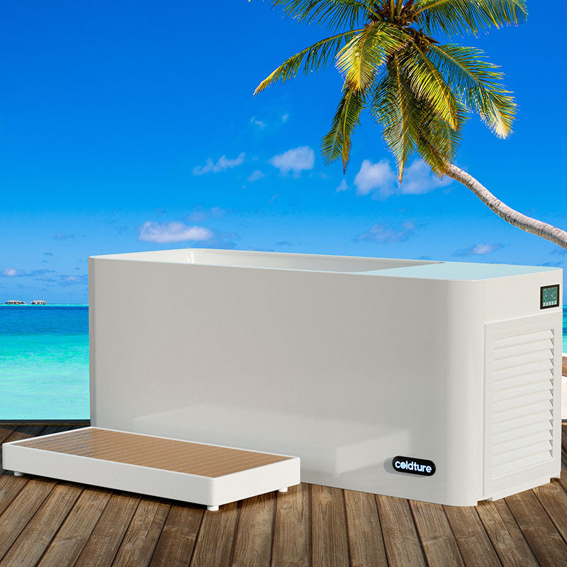 Coldture Pro Plunge, a sleek white plunge tub with a wooden step, set against a tropical beach backdrop featuring clear blue skies and palm trees.