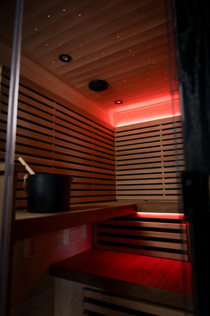 Infrared sauna interior with two-tier wooden benches and red LED lighting throughout the ceiling and walls. Black bucket and ladle sit on the upper bench.