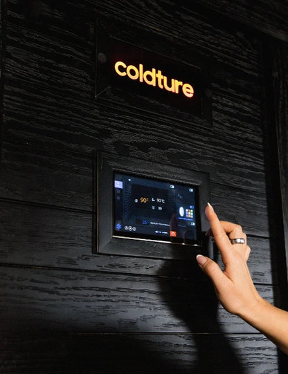 Hand adjusting a digital control panel on a black wood-clad sauna wall with yellow "Coldture" branding above, displaying temperature and heating settings.