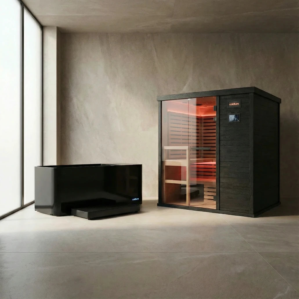 Black sauna cabin with glass front door and red interior heating elements illuminated, displayed alongside a separate black bench in a minimalist concrete room with large window.