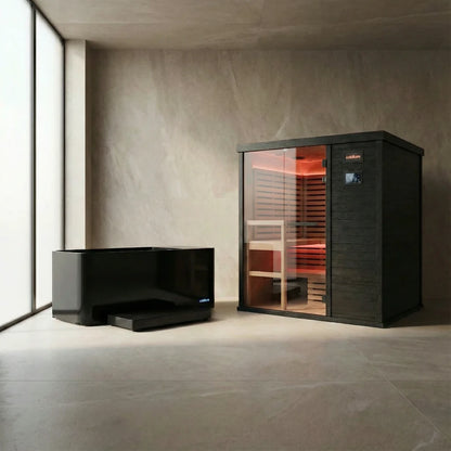 Black sauna cabin with glass front door and red interior heating elements illuminated, displayed alongside a separate black bench in a minimalist concrete room with large window.