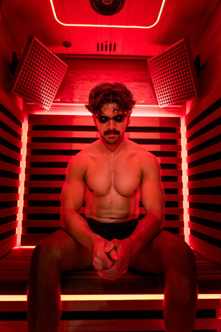 Man sitting inside a red light therapy sauna chamber with dual heat panels on either side, neon accents around the frame, and ventilation grilles visible at the top.